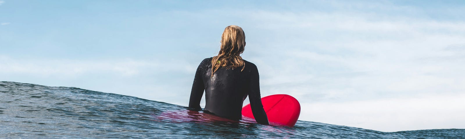 A female surfer clad in a wetsuit, sitting in the ocean on her red surfboard, waiting for waves