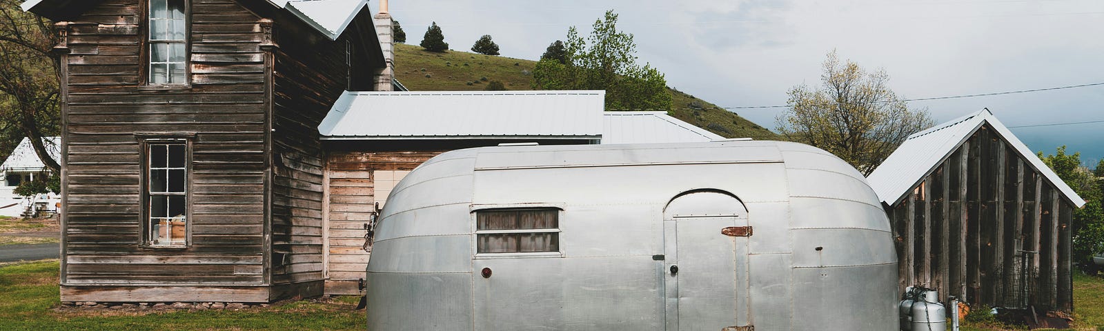 An old metal caravan, maybe a Windstream, from the 50s, parked behind abandoned buildings.