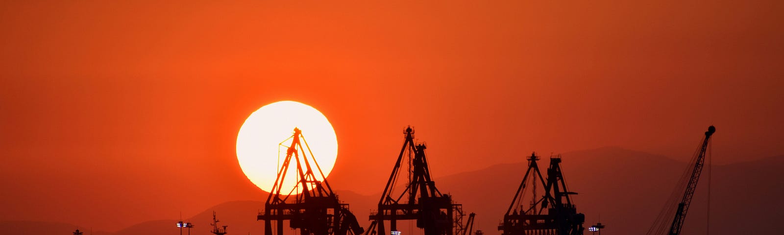 View from the sea of a port with various cranes in silhouette overlooking its harbour. The sun has begun to set behind the hills in the far distance, making the whole sky a vivid orange.