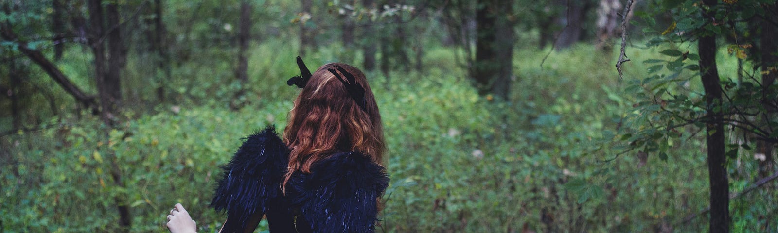 An unusual lady walks in a forest through tall seeds. Her hair is auburn and her dark dress features wings on the back.