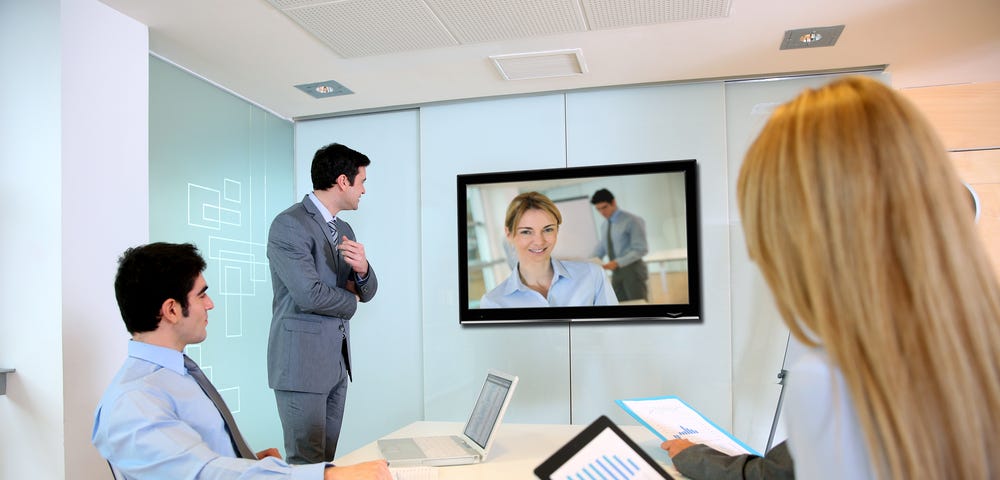 A group of two men and one woman surround a video screen where a video conference features a woman and a man