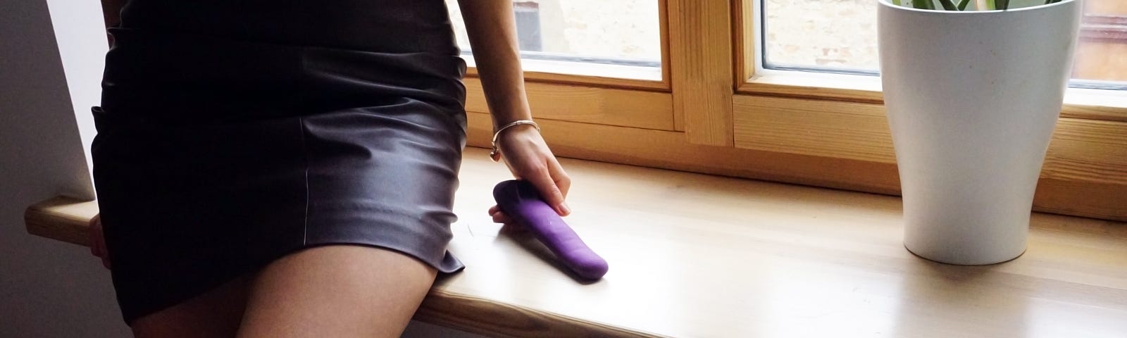 An image of a person in a short skirt, standing with her back to a window, leaning against the window sill and holding what looks like a sex toy in her hand.