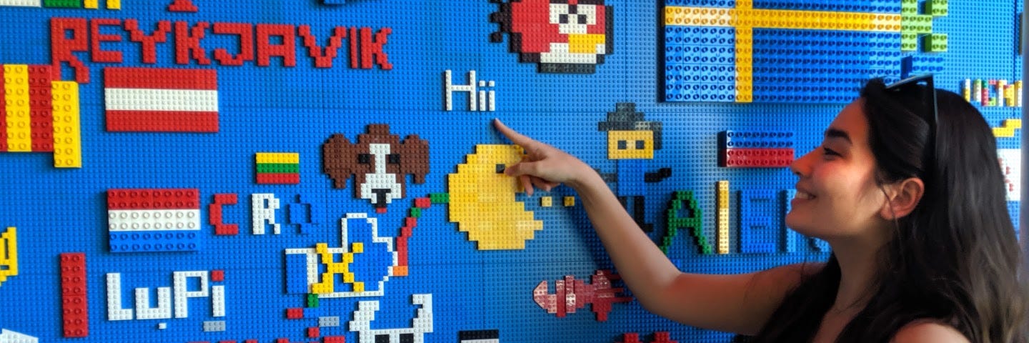 A person with long black hair smiles and points at a multi-colored wall made of legos.