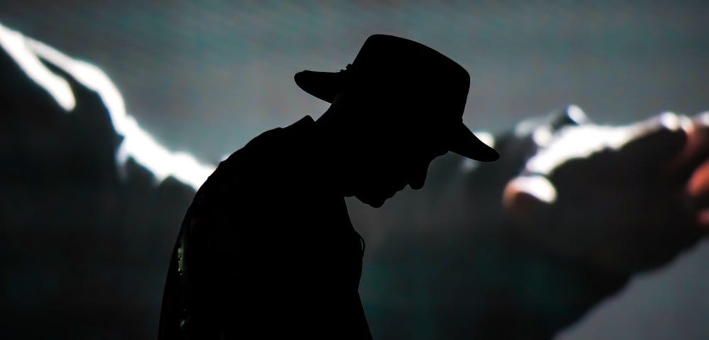 The silhouette of a man wearing a cowboy hat/fedora, bowing slightly. Behind him, a projection of an arm (a cinema, perhaps).