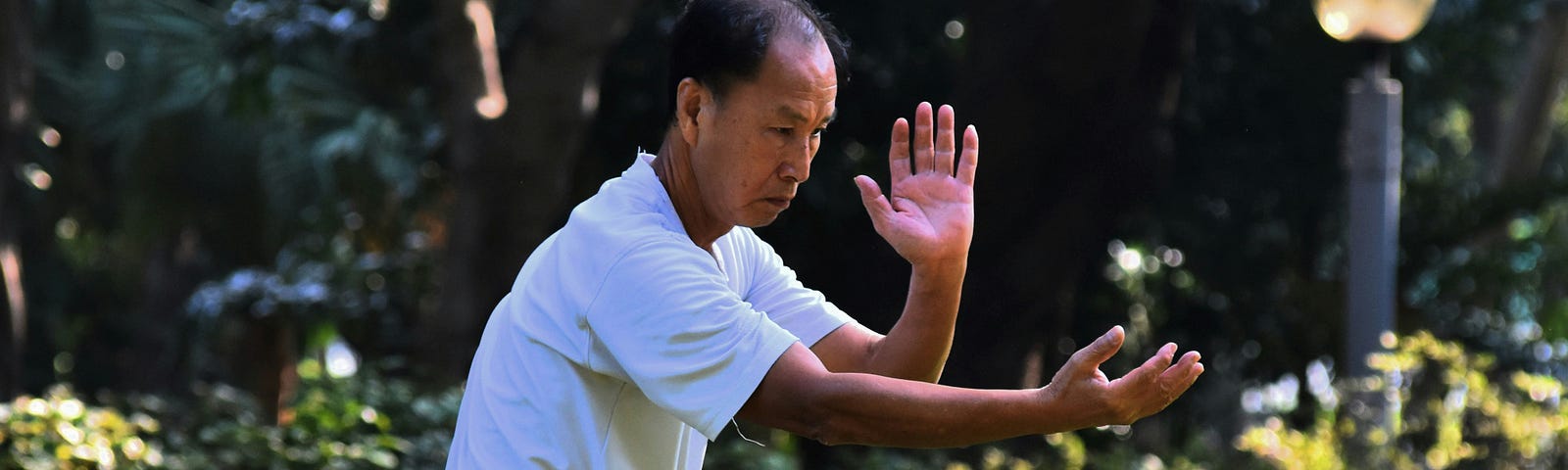 A coloured image showing a man wearing a white teeshirt and black jeans practicing Tai Chi in a park.
