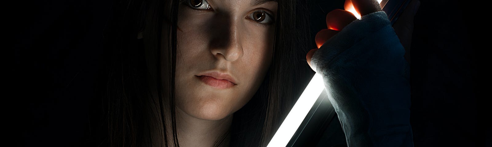 Photo of a woman's face staring down the camera, as she's gripping a sort of light sabre.