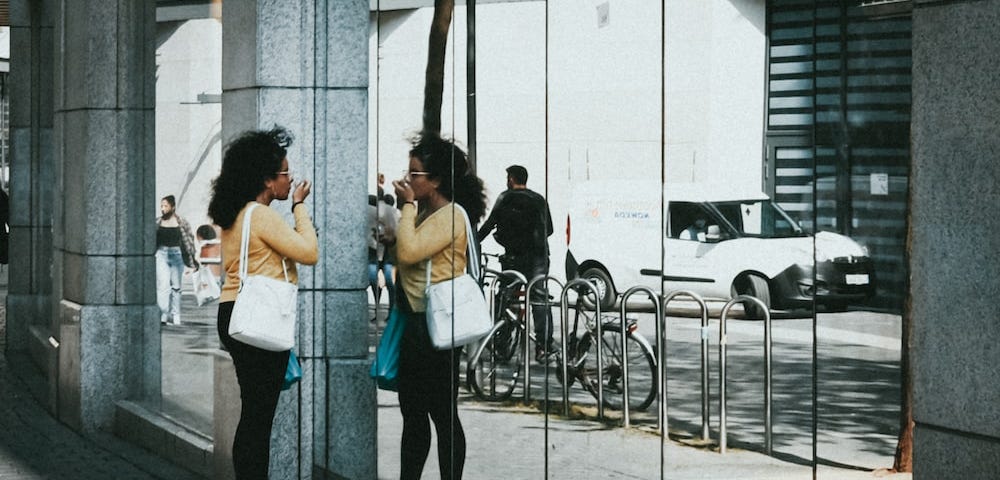 Young lady admiring herself in a window. Wearing a yellow sweater, blue jeans, white shoes with a white bag on her shoulder.