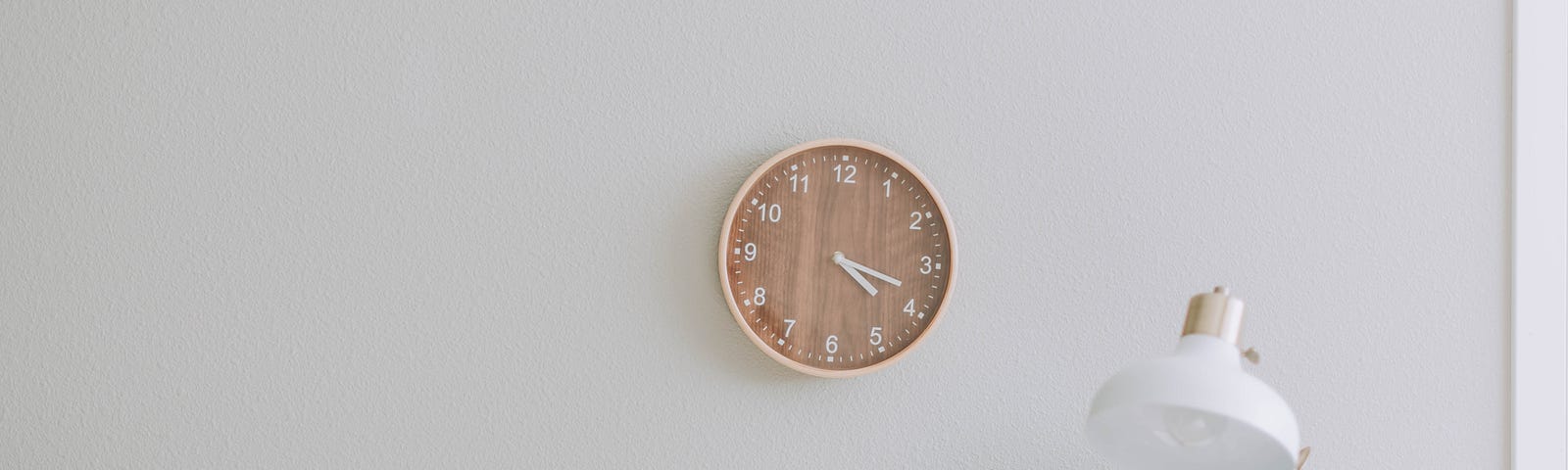A minimalist composition featuring a plain grey wall as the backdrop. In the foreground, a wooden digital clock is displayed alongside a simple white lamp. On a white table beneath them, a plain broad-leafed plant in a white bowl adds a touch of greenery to the scene.