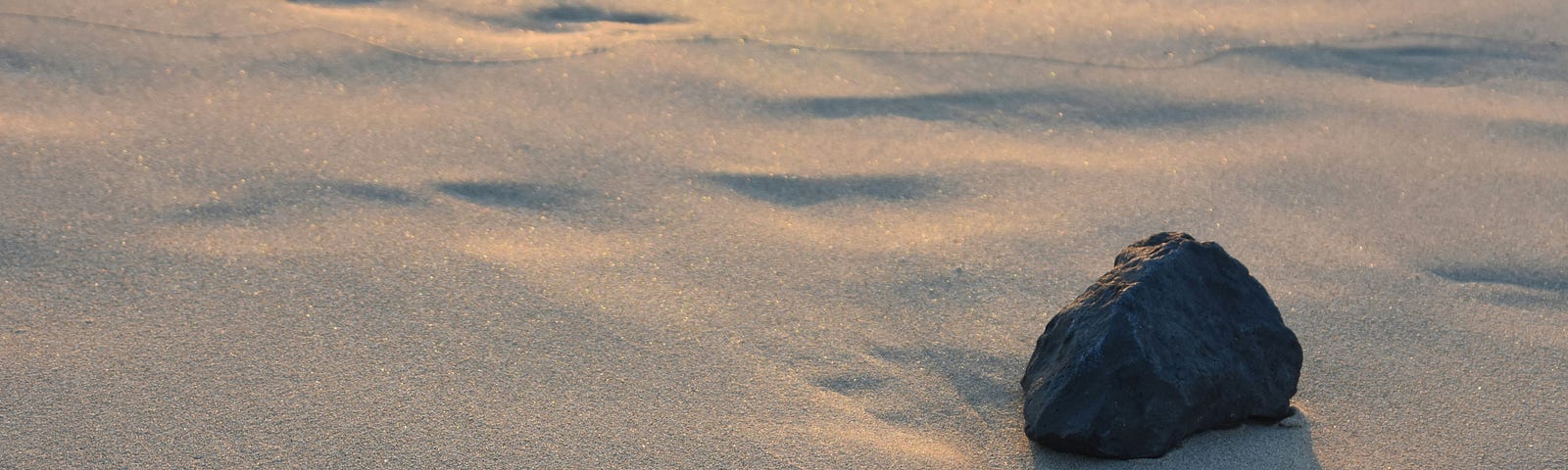 sand on a beach with scattered imprints, a black rock sits, shaded with bits of light