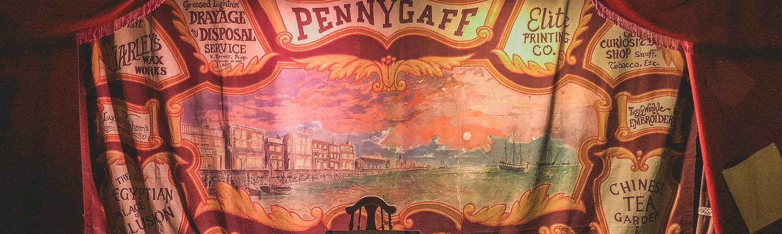 An empty stage with a high-back wooden chair in the middle. There is a backdrop in reds, golds mostly that says Pennygaff at the top. This was a type of entertainment for the lower classes in 19th century England. There is a river scene with tall masted ships in the river.