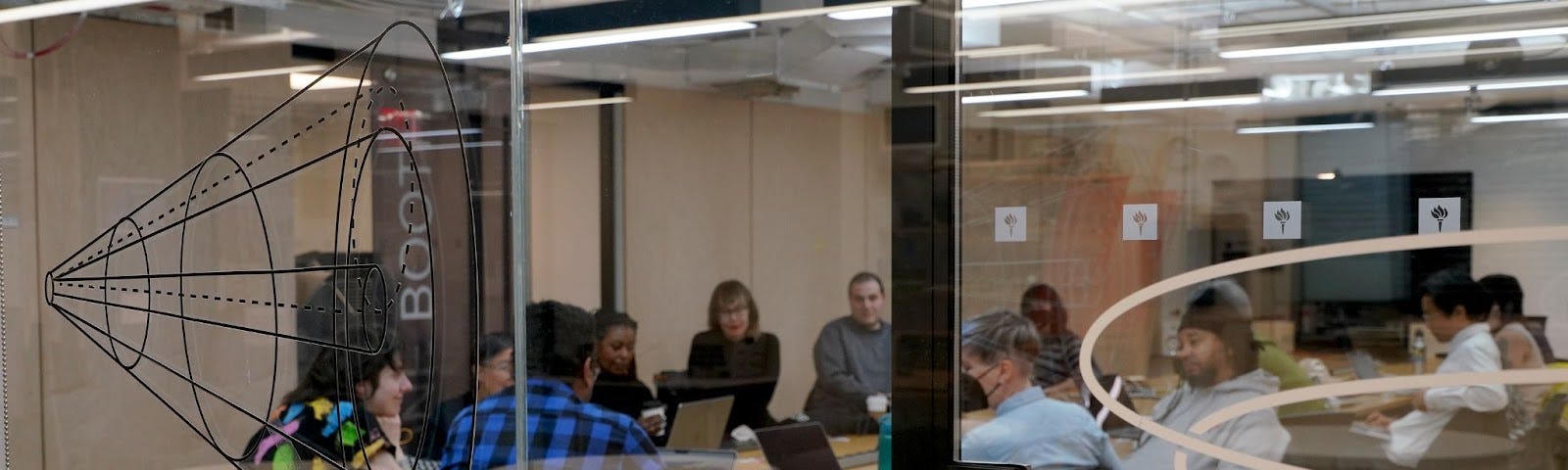 A behind-the-scenes image of our board of directors and advisors, staff, and community members at NYU ITP. The image is taken in front of a glass door. Inside the room, around 10 people gathered by a table, working on their laptops.