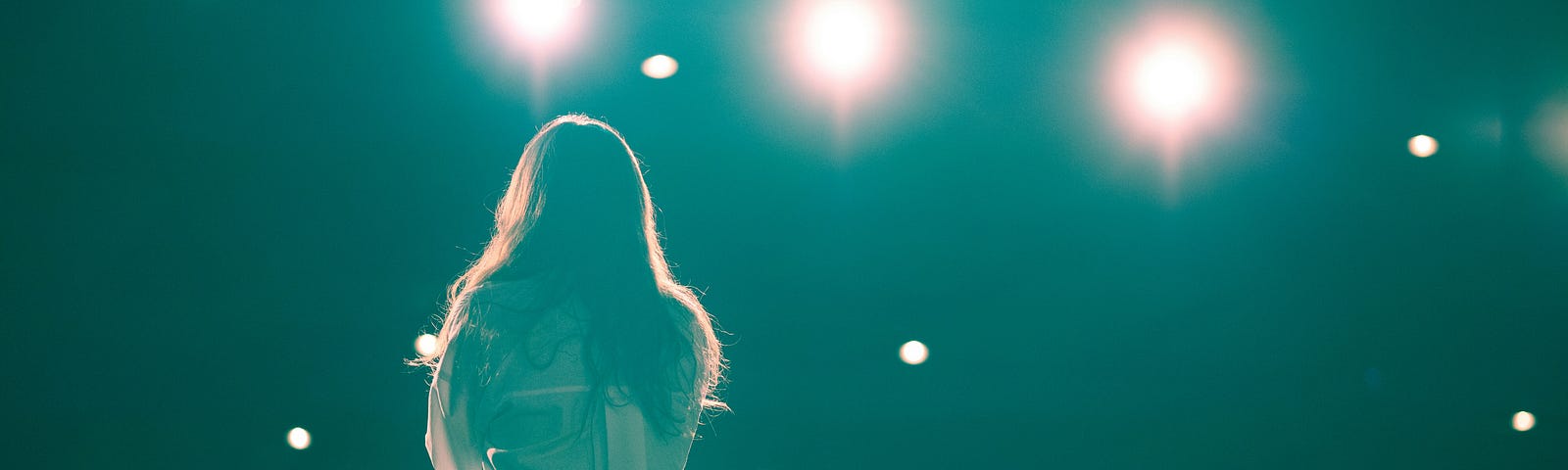 Woman on stage with back to camera, multiple spotlights lighting up in front of her. The picture is washed in a greenish-black tone.