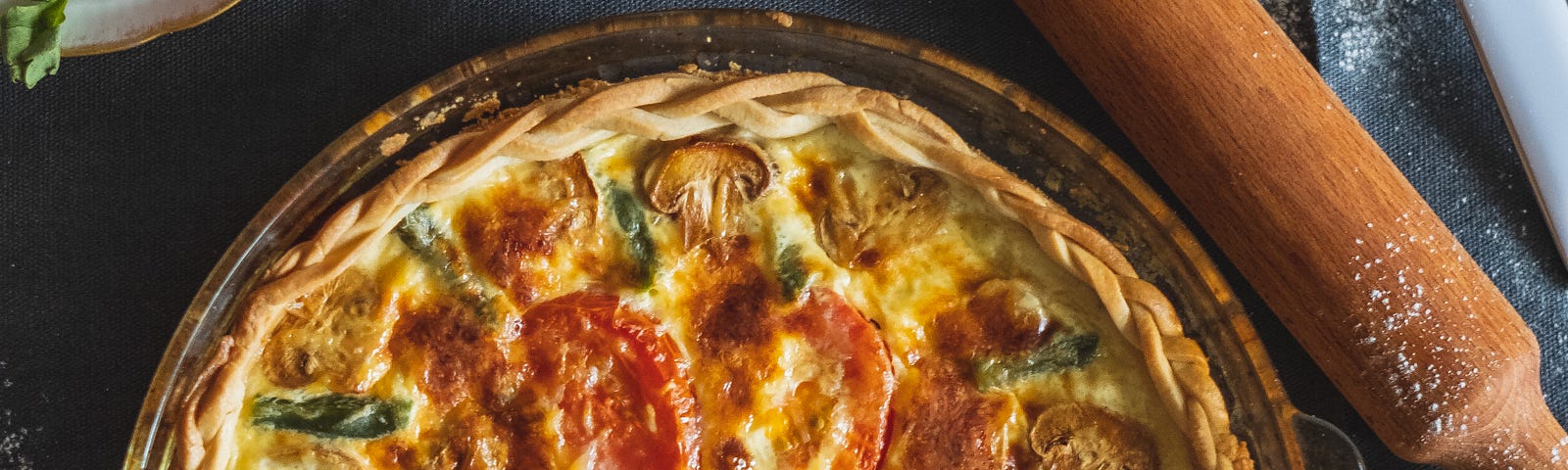 A quiche with tomatoes mushrooms and sugar snap peas, on a table with a rolling pin and a wooden spoon.