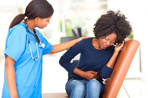 a Black female doctor with her hand on the shoulder of a young,Black woman who is grimacing with abdominal pain