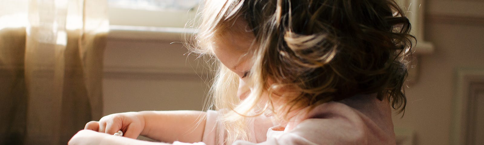 A side-view of a little girl, barely the height of the windowsill, wearing a pink dress holding out a string of pearls as if determining to put them around her neck. Her hair short and wavy but maturely coiffed like a woman’s but with a barrette. A representative photo of a girl growing up too fast against more idealistic fashion.