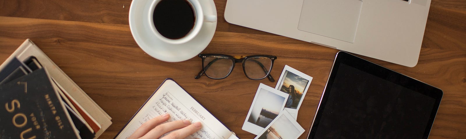 A person writing something in a journal notebook on a desk with a laptop, cup of coffee, photos, and a stack of books.