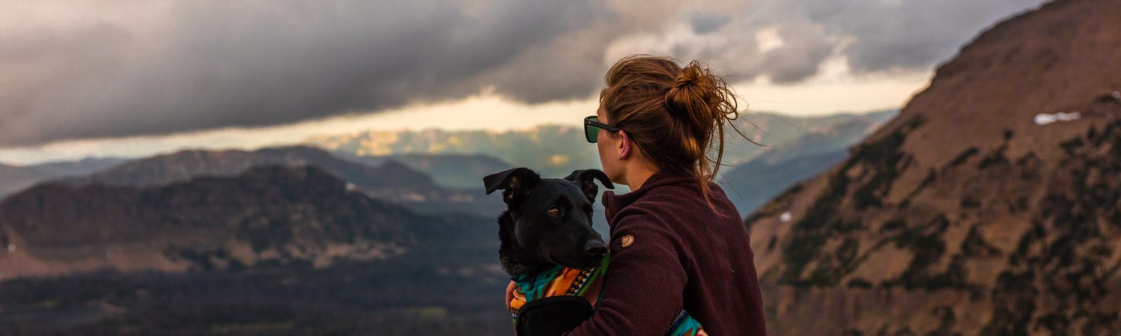 A woman hugging her dog and watching a storm roll in while her dog checks for activity behind them.