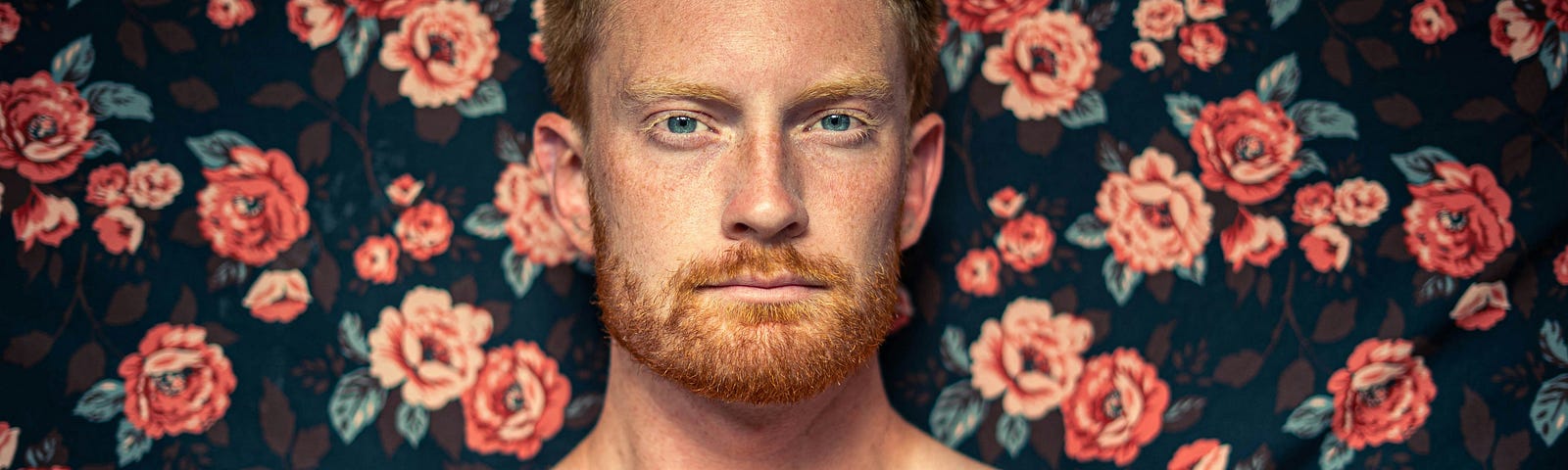 A man with freckles poses in front of a flowery wallpaper backdrop.