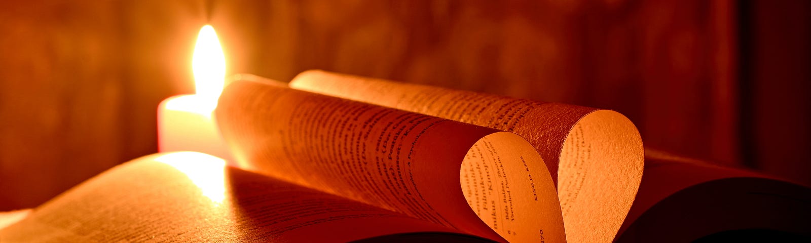 A Bible-looking book is lays open near a candle. two pages are folded into ht center forming a heart shape when seen from the bottom or top. the heart glows golden in the candlelight. but it could be a dictionary!