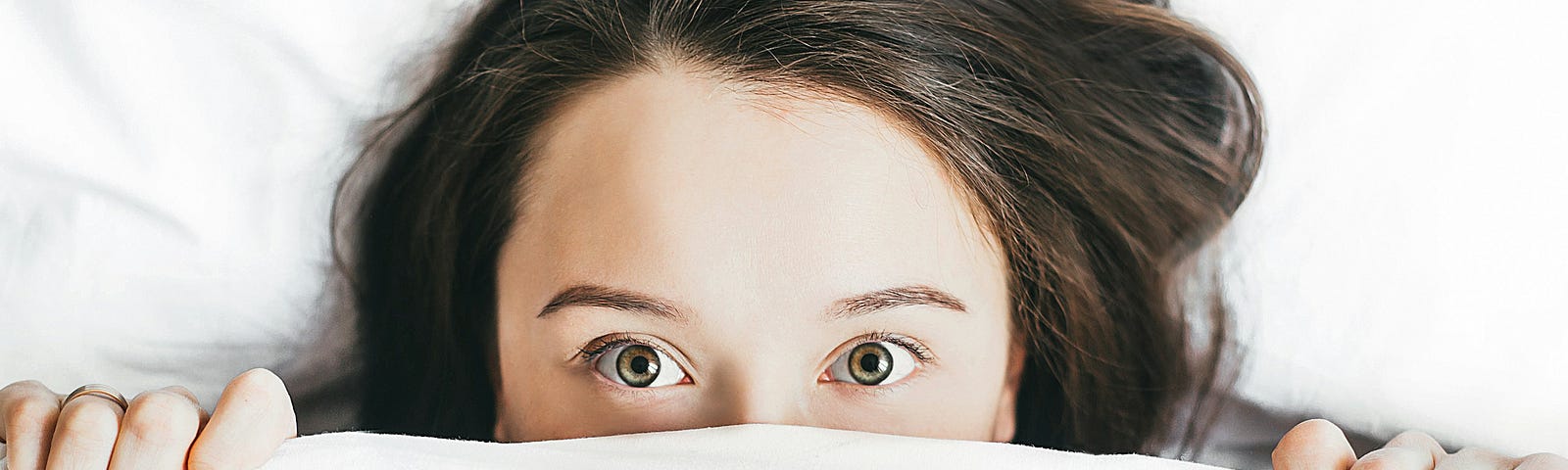 A woman with the bed covers pulled up only exposing her eyes and top of her head.