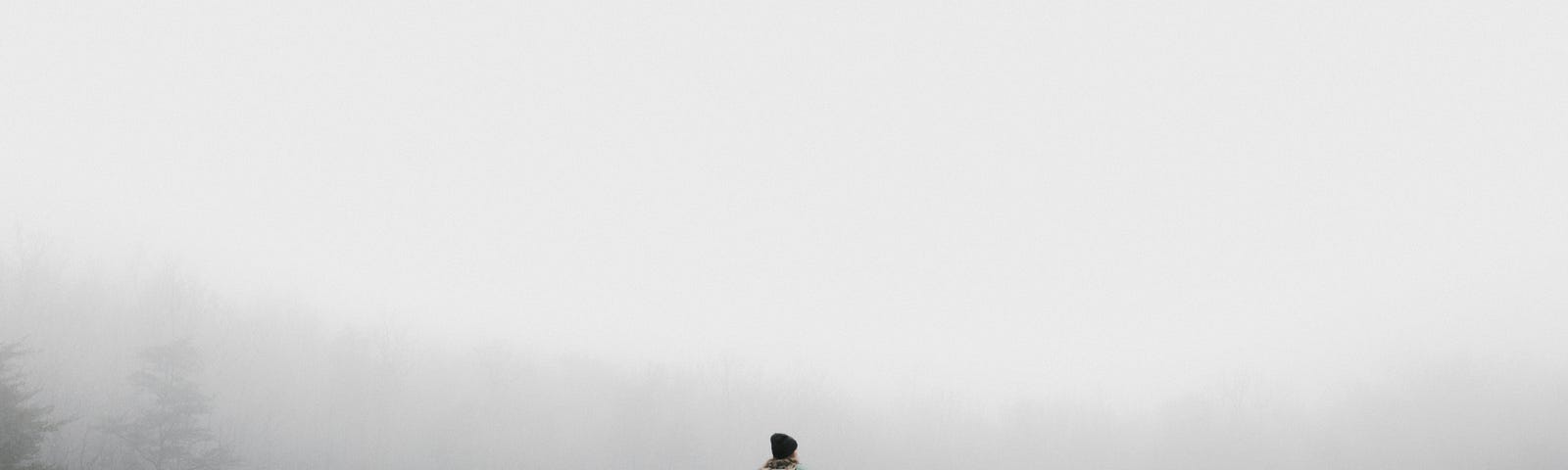 A backpacker stands in front of a forest shrouded in fog