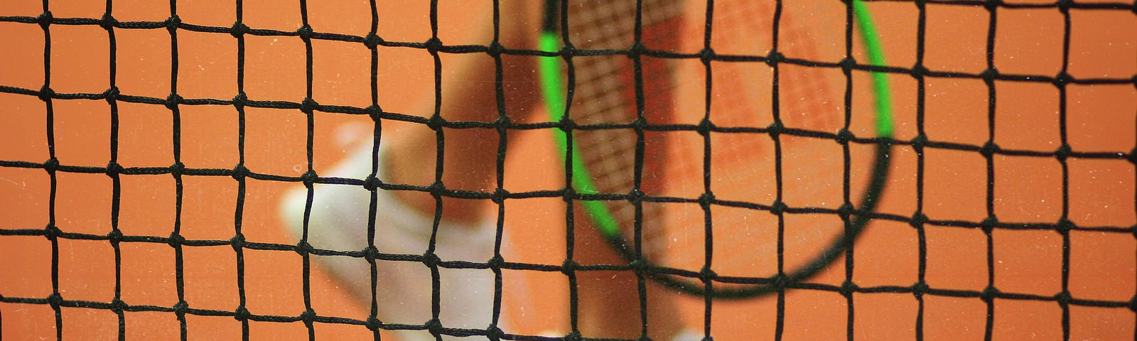 A photograph of the legs of a tennis player holding a tennis racket next to a tennis ball through a net.