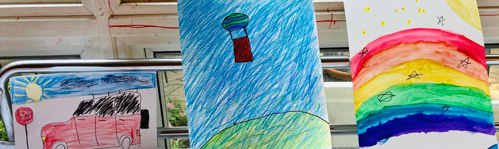 Picture by Author. Drawing of a car. Drawing of a hot air balloon. Drawing of a rainbow. All three pictures are sitting on a bed up agaisnt the window on a blanket with red hearts. My adult son is sleeping and is not in picture.