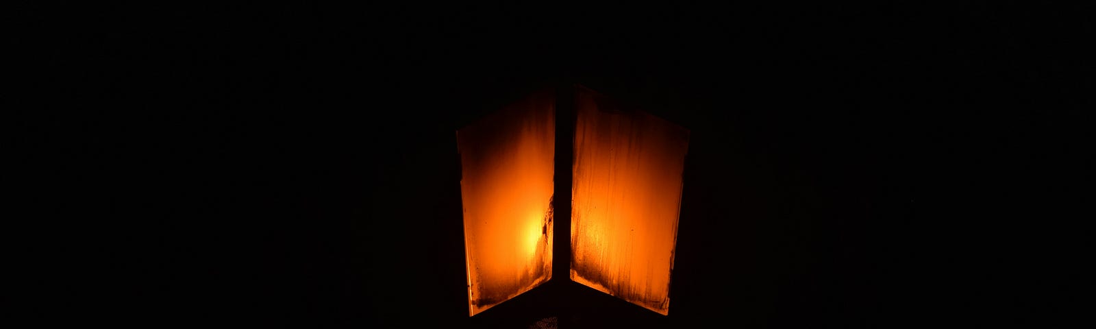 A canted view from below of what looks like a gas streetlamp. It glows orange is enveloped in a completely black background.