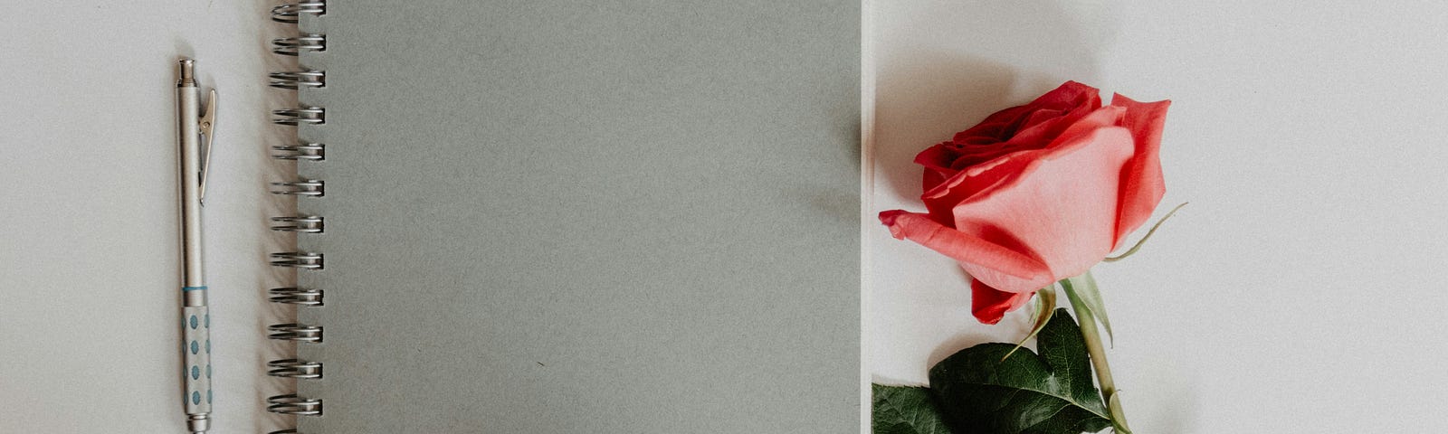 A closed notebook with a pen and a pink rose next to it.