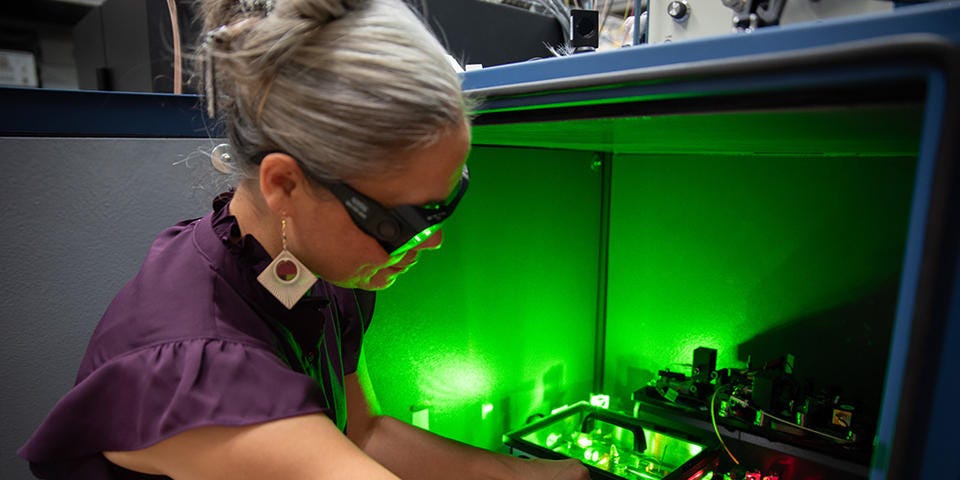 A researcher wearing safety glasses reaches into a box of circuitry and other equipment, which emits a green glow.