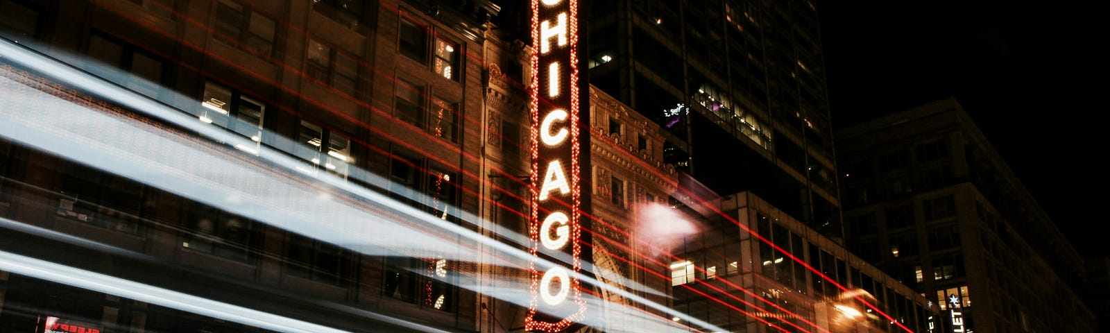 Streaks of car lights fo the lenfth of a city street, with a theater sign (reading “Chicago”) looming large in the background.