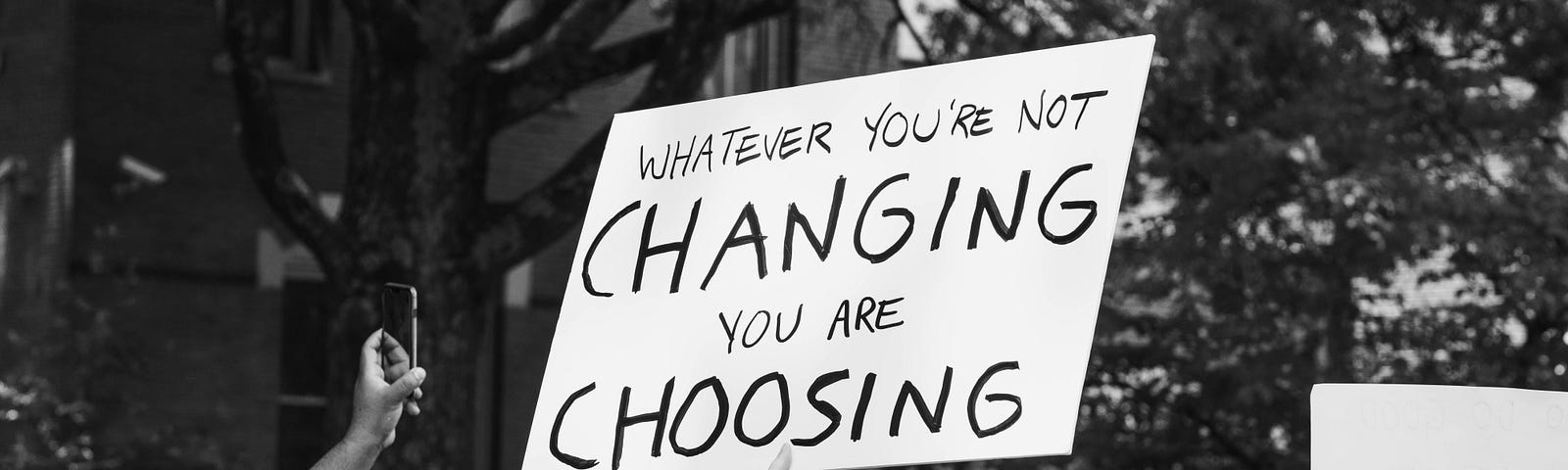 A protestor holding a sign that says “Whatever You Are Not Changing You Are Choosing”