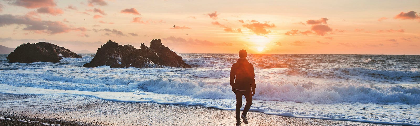 A clothed man (seen from behind) walks towards incoming waves at the beach as the sun begins to set. Large rock outcrops surge upwards out of the sea in the midground at the left.