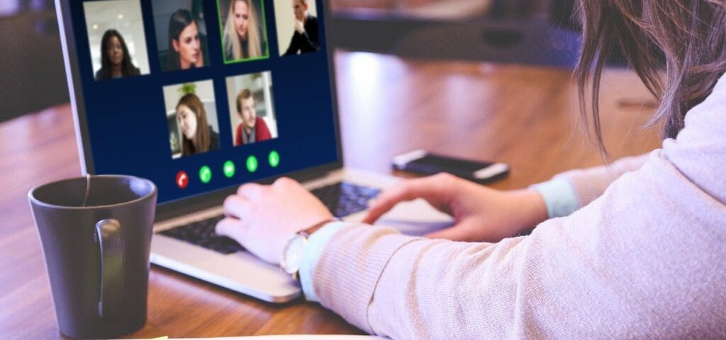 Photograph of a woman at a table using a laptop to take part in a video call, with a notebook in the foreground