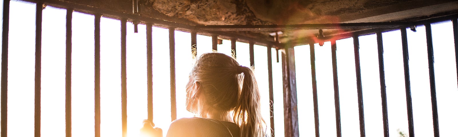 Woman looking out from behind bars, sun shining in on her.