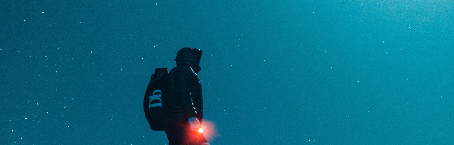 A man on a cliff looking at the night sky witha red glowing beacon in his hand