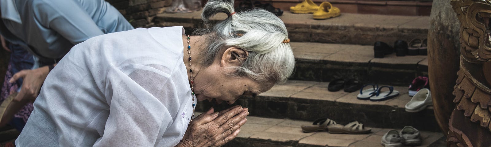 An elderly woman bowing with hands clasped in prayer outside a place of worship