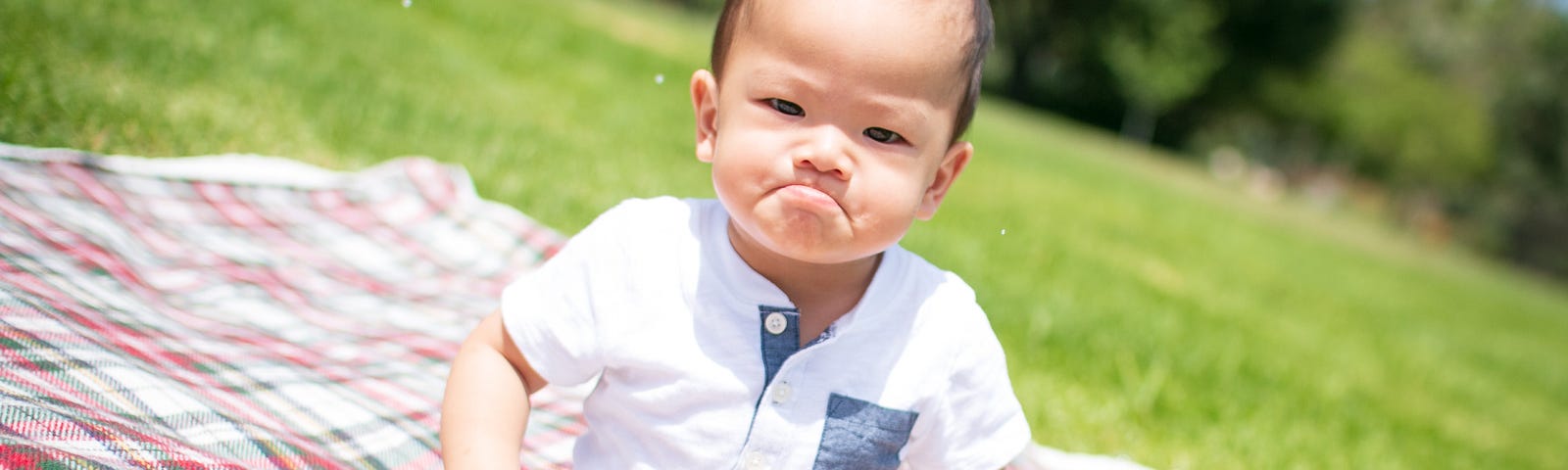 Asian baby sitting on a plaid blanket over grass. He is ostensibly frowning.