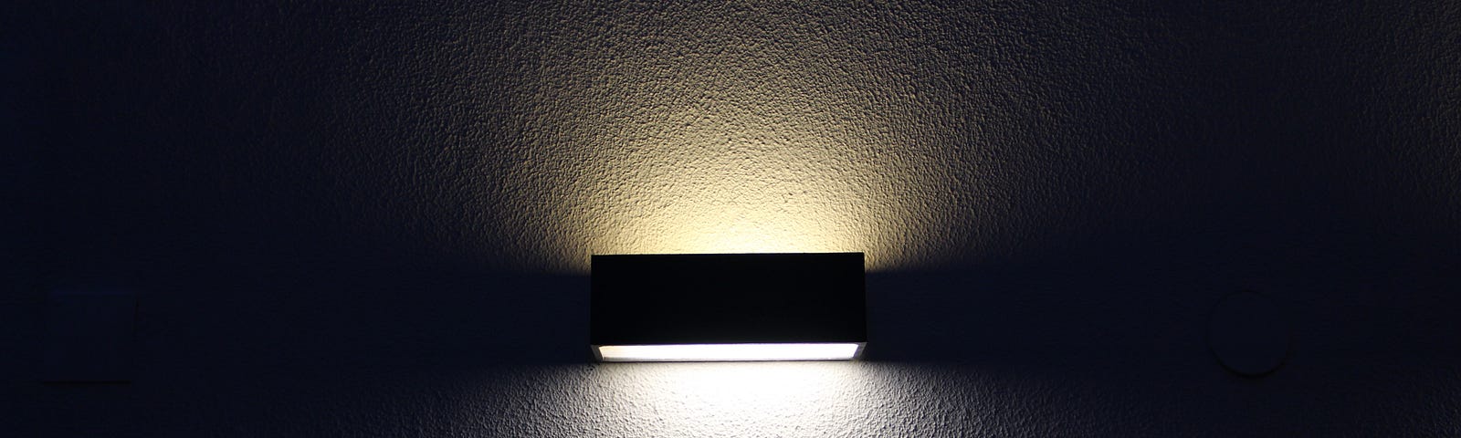 A solitary rectangular light casts a bright glow and a shadow across a wall