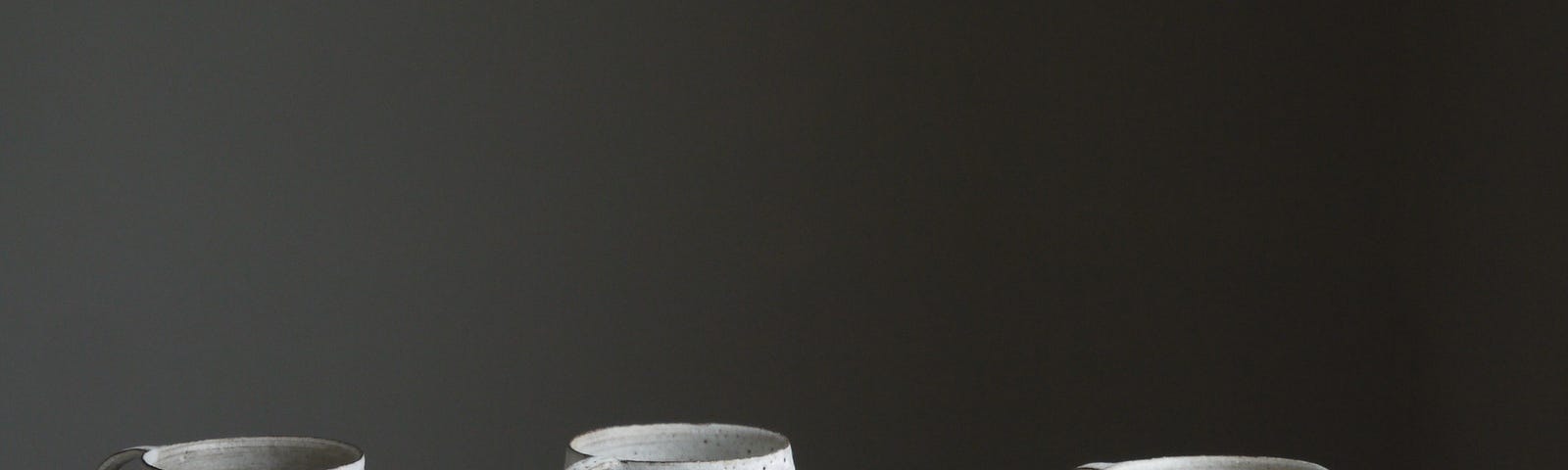 White mugs on a shiny white table, against a black background. The mugs have irregular shapes.