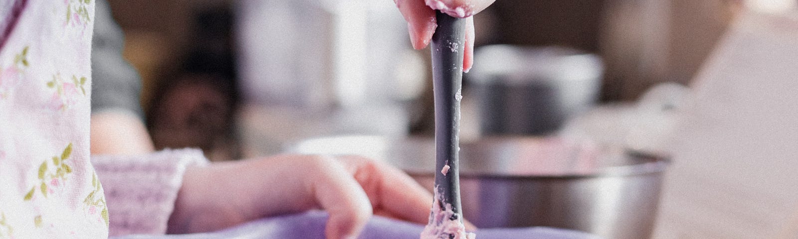 A woman standing over a purple mixing bowl with a spoon mixing pink icing.