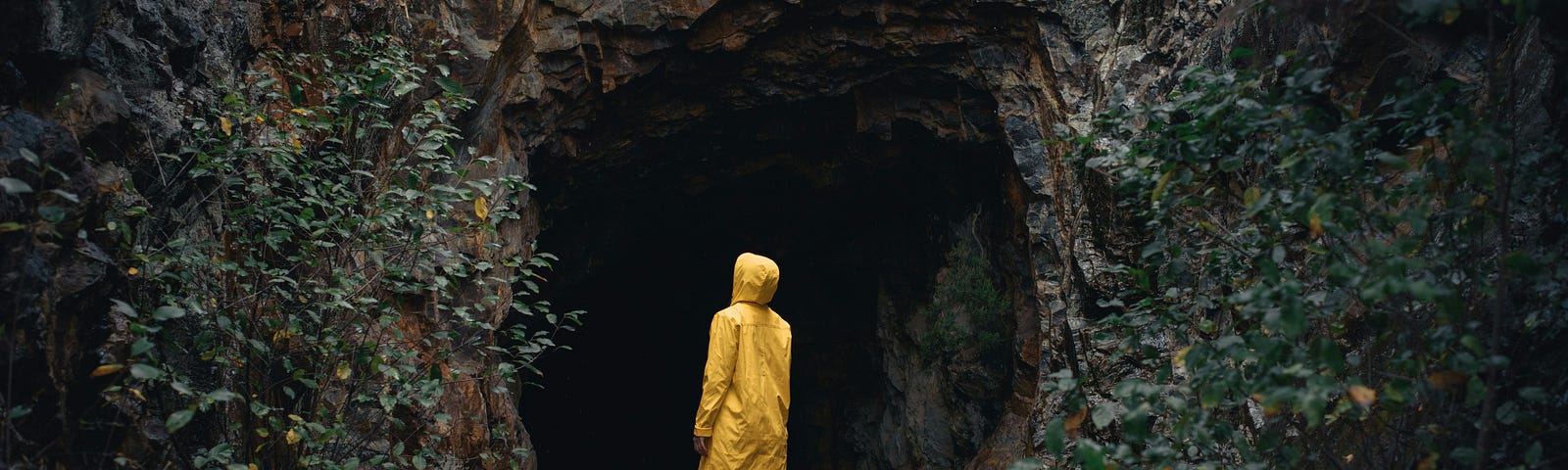 A woman in a strong yellow raincoat standing at the entrance of a large cave looking at its darkness. We all go though dark moments. But can come out strengthened.