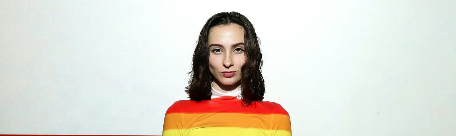 A woman stands in front of a rainbow backdrop with rainbows on her shirt.