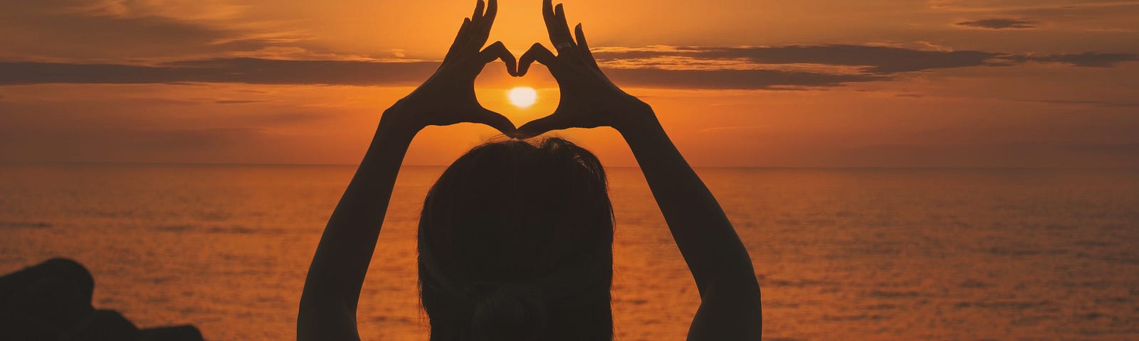 A woman watches a sunset at the beach and forms a heart with her hands.