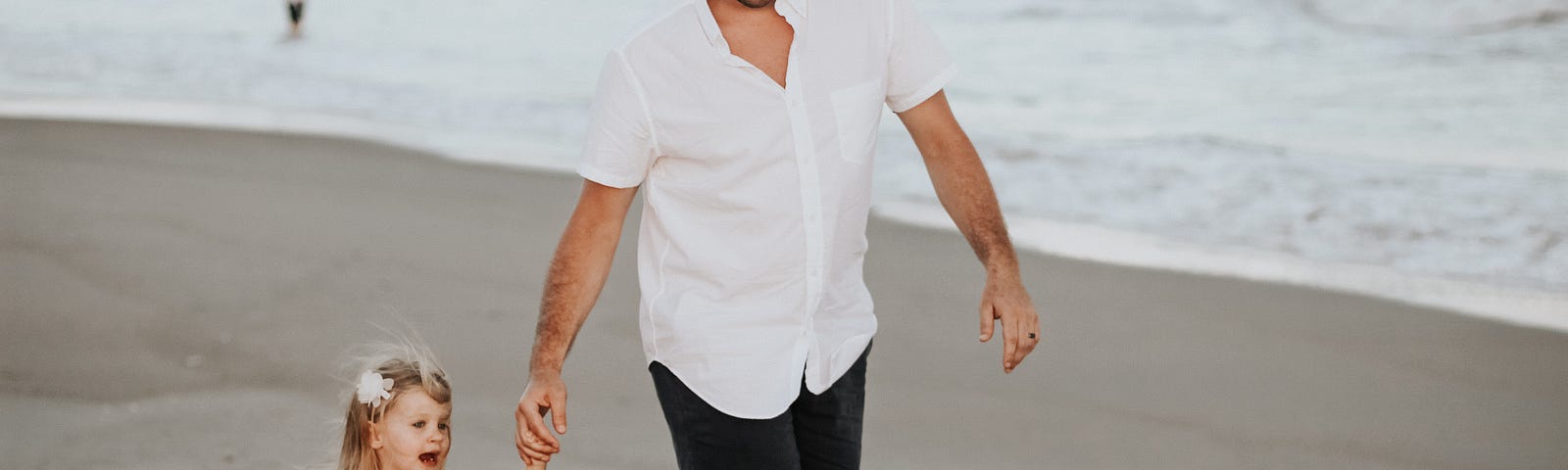 Father and little girl holding hands walking on sand with ocean in background