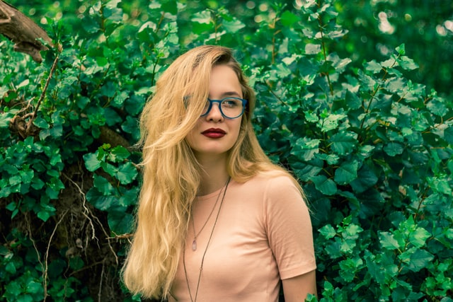 Attractive Young Blonde Woman Wearing Glasses And Red Lipstick Standing Next To Green Shrubs In A Field.