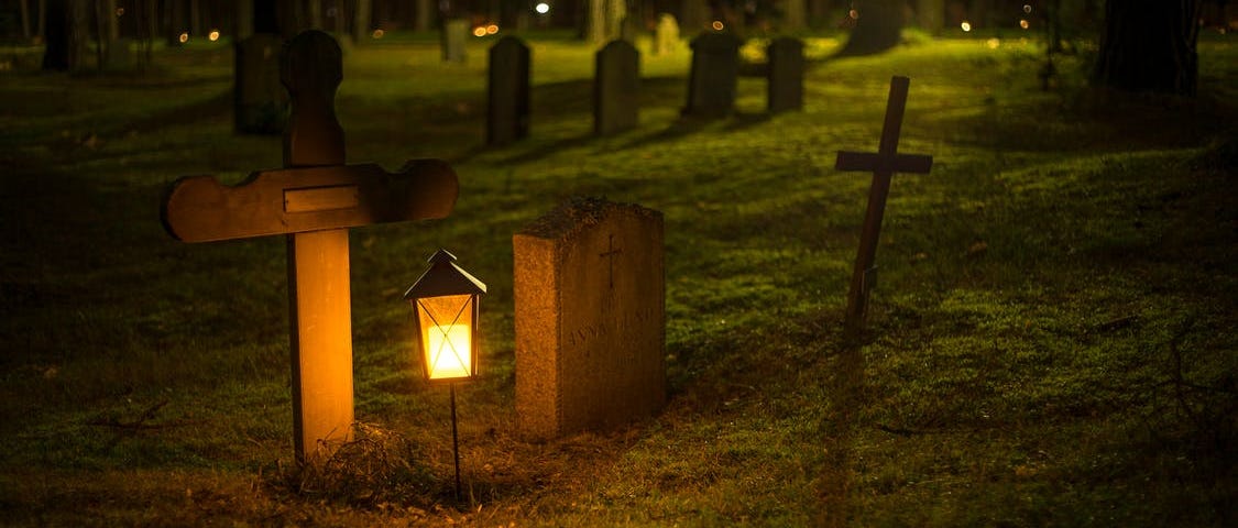 An image of several gravestones in a graveyard at night. A lantern glows gold in the foreground and lights the stones.