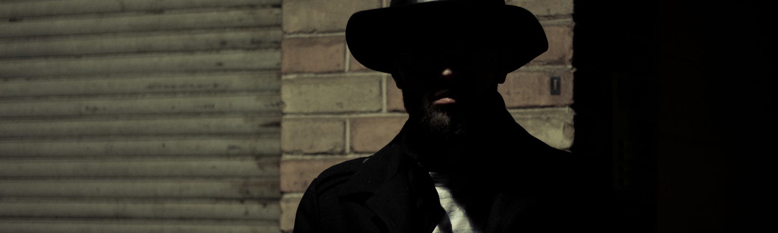 Man in a dark suit and fedora with his face hidden in shadows.