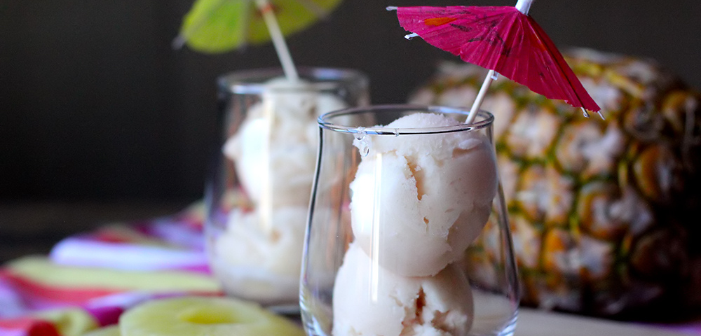 A photograph of two clear glasses with two scoops of a white frozen pineapple and guava sorbet. They have decorations of a cocktail umbrella in each dessert, one is dark pink and the other lime green placed in the first scoop. A fresh pineapple is behind the frozen desserts and in front are pineapple slices and a spoon. Everything is placed on a cutting board with a colorful cloth underneath.