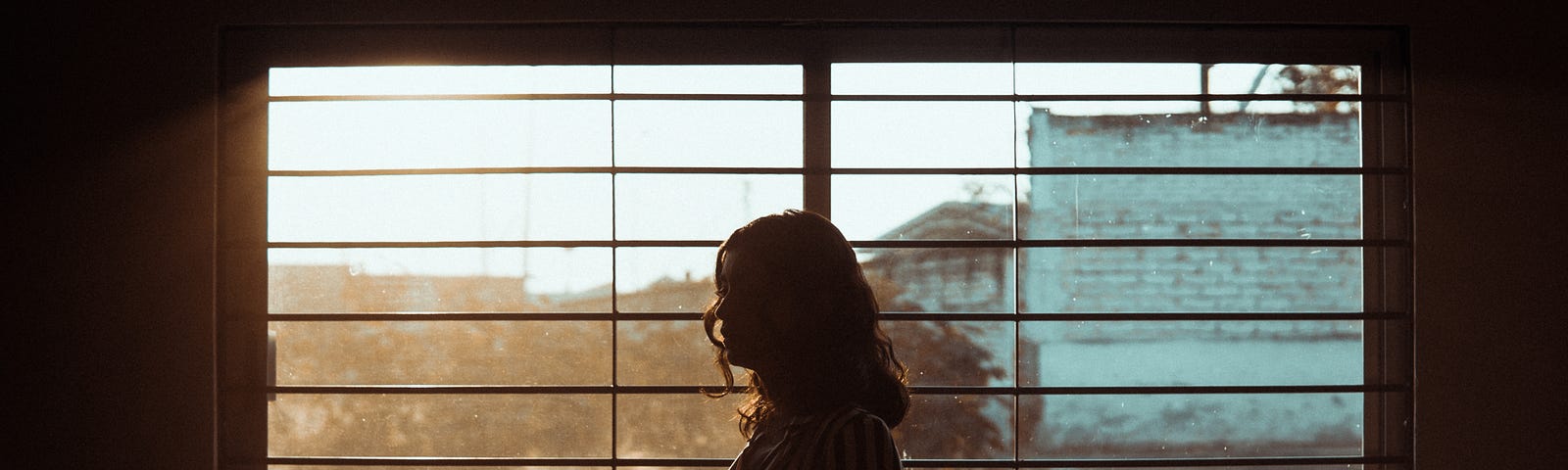 A woman looks out at buildings beyond a window, her body silhouetted against the light.